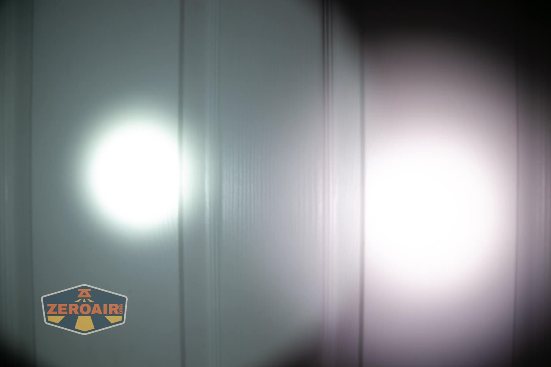 Two flashlight beams shine side by side on a white vertical panel wall, creating overlapping circular areas of light with different brightness and color temperatures. A ZeroAir logo is visible in the lower left corner.