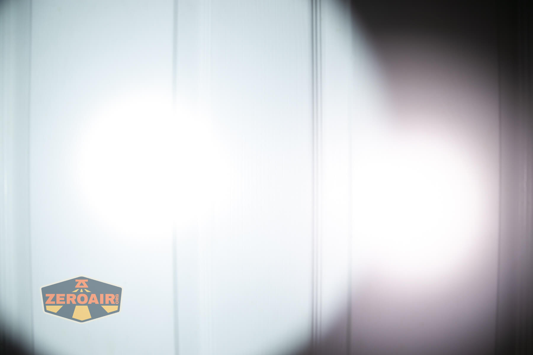 A bright white flashlight beam and a dimmer round spot are projected onto a white paneled door. The ZeroAir logo appears in the lower left corner.