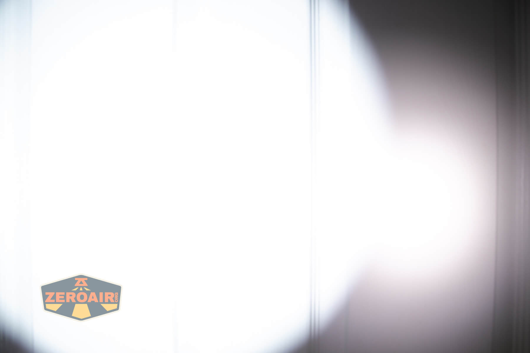 A bright white light is shining on a wall, creating a circular beam pattern. The background is mostly white from the intensity of the light. A small ZeroAir logo appears in the lower left corner.