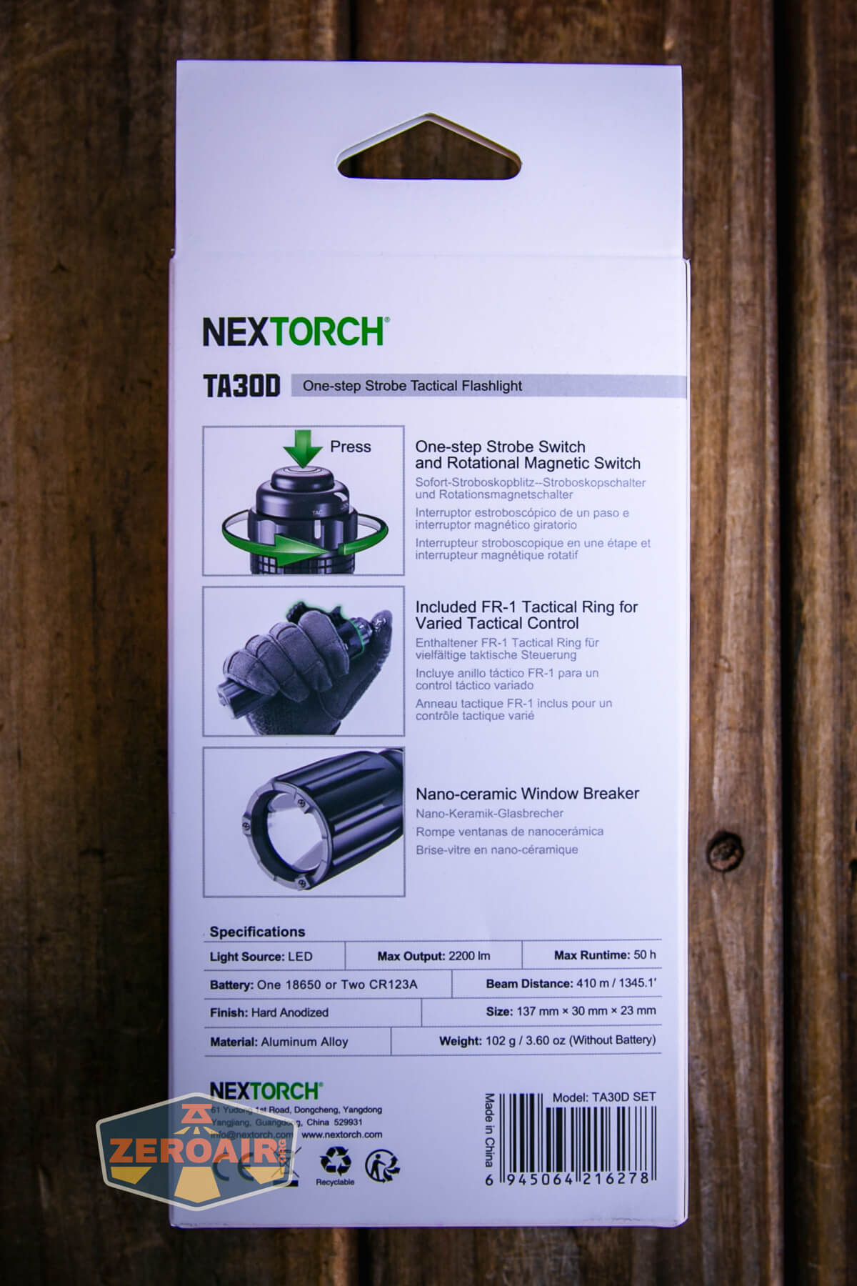 Back of a NEXTORCH TA30D flashlight package showing product features, specifications, and images of the flashlight, tactical ring, and window breaker. The packaging sits on a wooden surface.