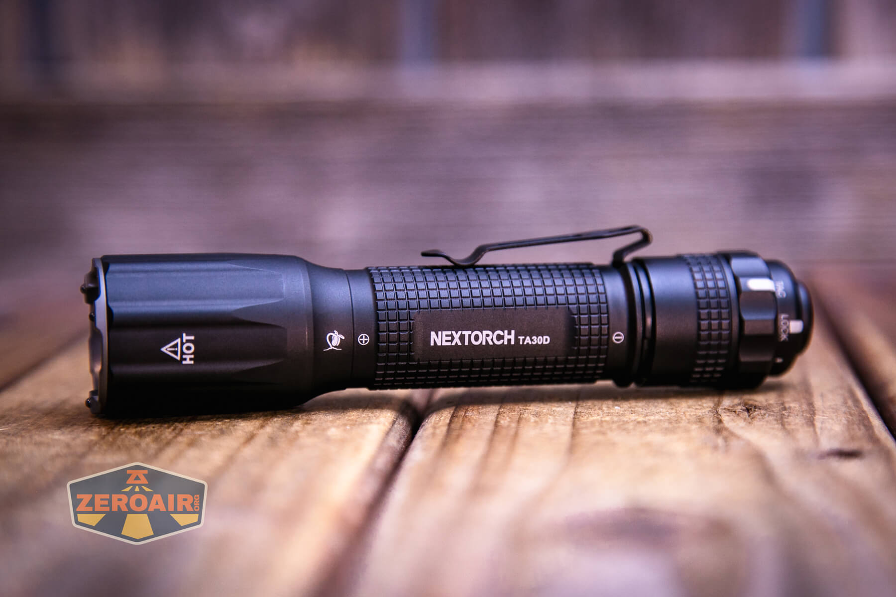 A black NEXTORCH TA30D flashlight with a textured grip and clip lies on a wooden surface. A “ZEROAIR” logo appears in the lower left corner of the image.