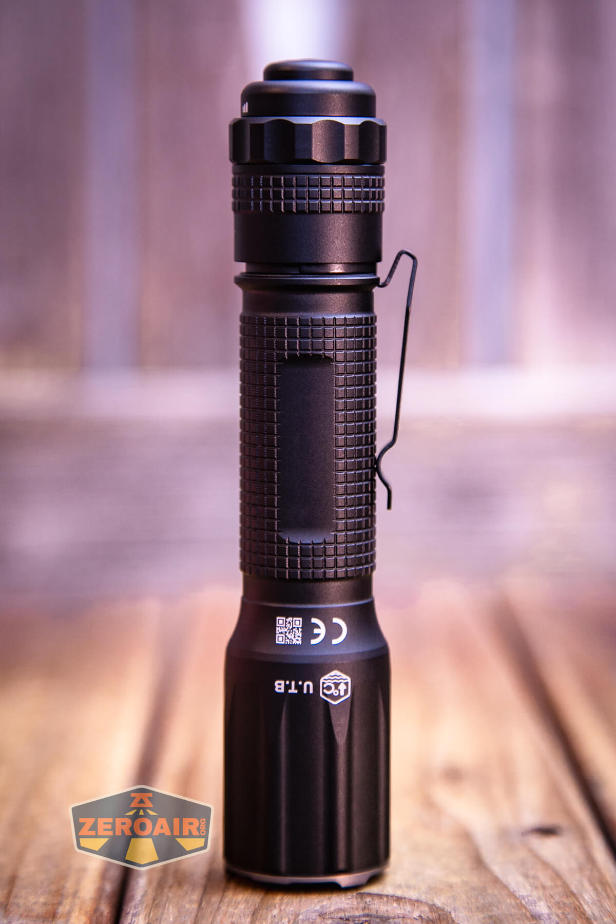 A black handheld flashlight with a textured grip and side clip stands upright on a wooden surface, with a blurred background and a ZeroAir logo in the lower left corner.