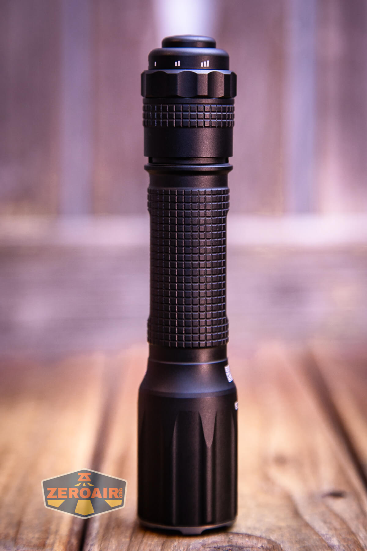 A black flashlight with a textured grip stands upright on a wooden surface, with a ZeroAir logo in the lower left corner. The background is softly blurred in shades of brown and gray.