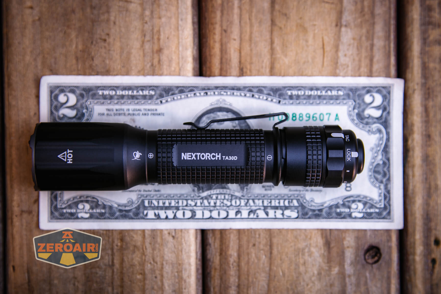 A black NEXTORCH TA30 flashlight is placed horizontally on a U.S. two-dollar bill, both resting on a wooden surface. The ZeroAir logo is visible in the bottom left corner of the image.