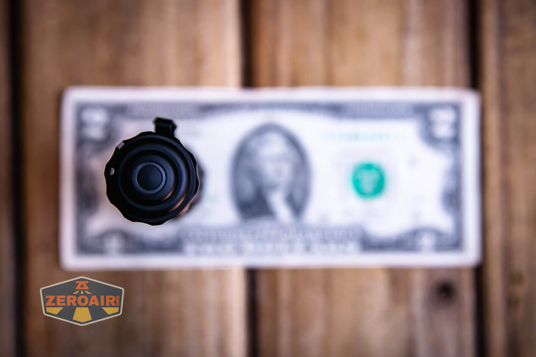 A small flashlight is positioned vertically, viewed from above, in front of a US two-dollar bill placed on a wooden surface. The image is slightly out of focus except for the flashlight’s button. A ZeroAir logo is in the lower left corner.
