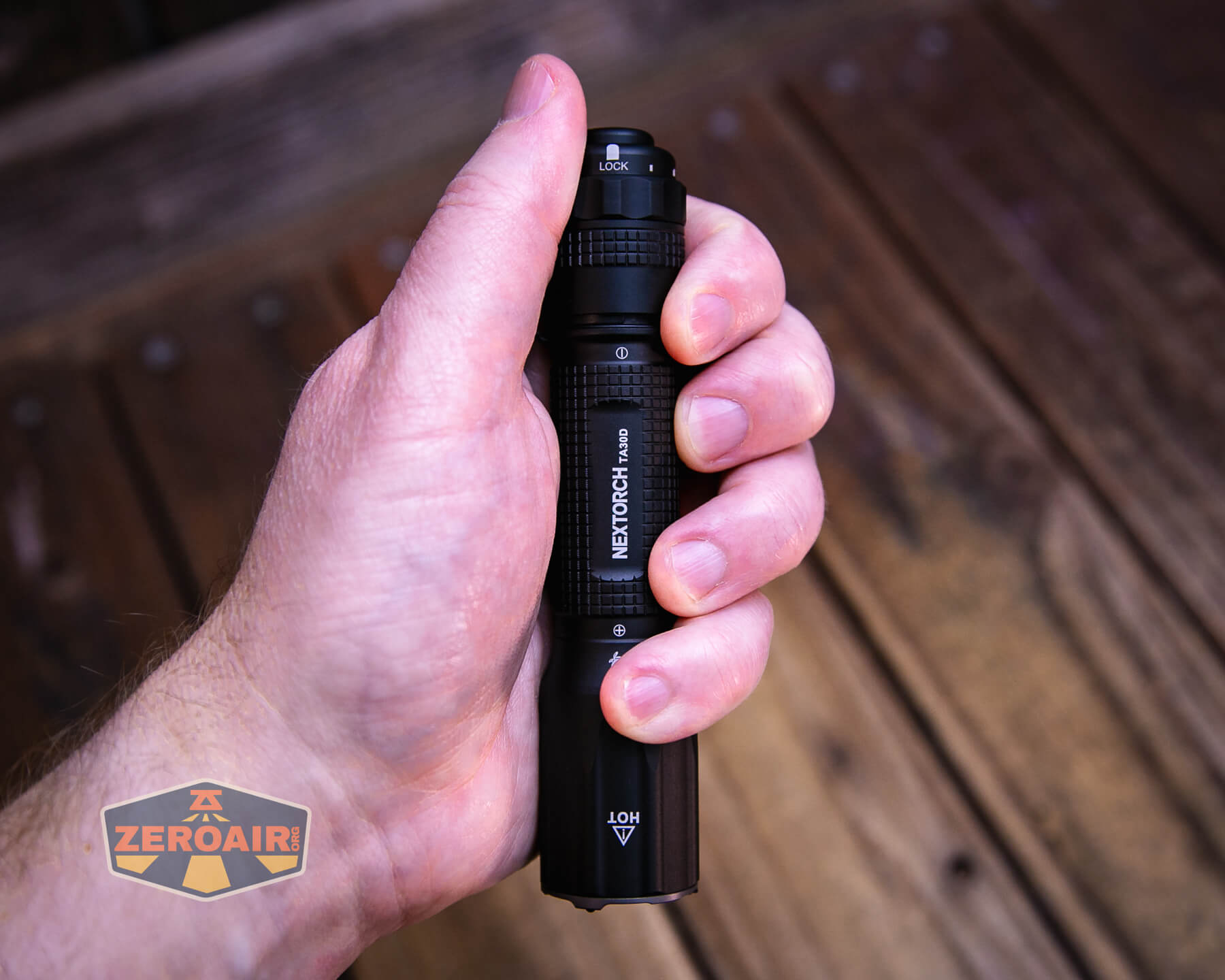 A hand holding a black NEXTORCH E52C flashlight with textured grip, pointed forward. The background shows wooden boards and a ZeroAir logo in the bottom left corner.