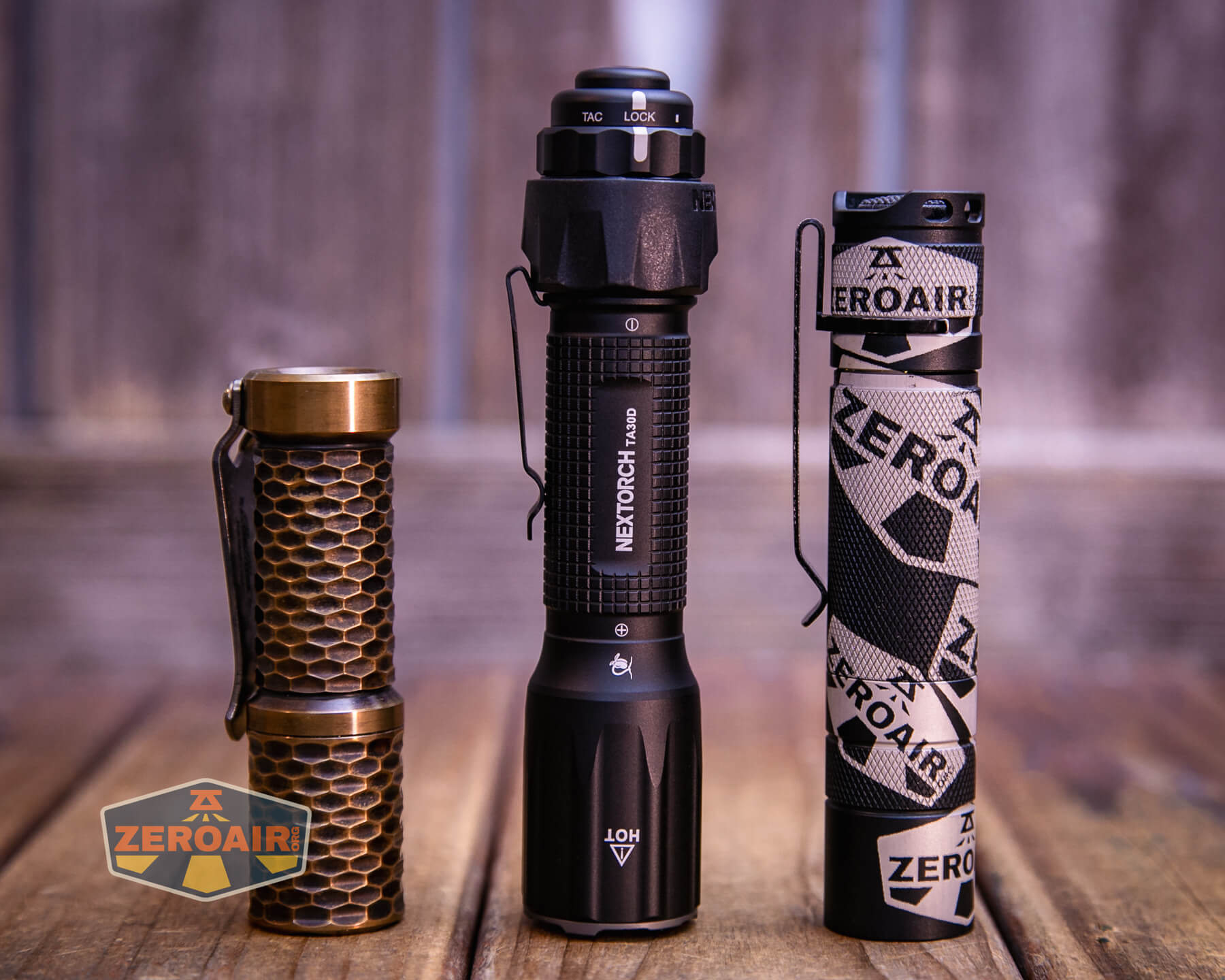 Three flashlights stand upright on a wooden surface. The left one has a hexagonal pattern and bronze color, the middle is black with textured grip, and the right has a patterned design with ZEROAIR text.