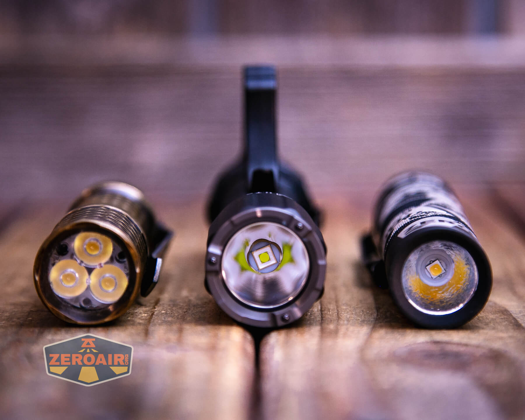 Three flashlights are placed side by side on a wooden surface, viewed from the front to show their LED bulbs. The left flashlight has three LEDs, the center has one, and the right has a single LED with a different design.