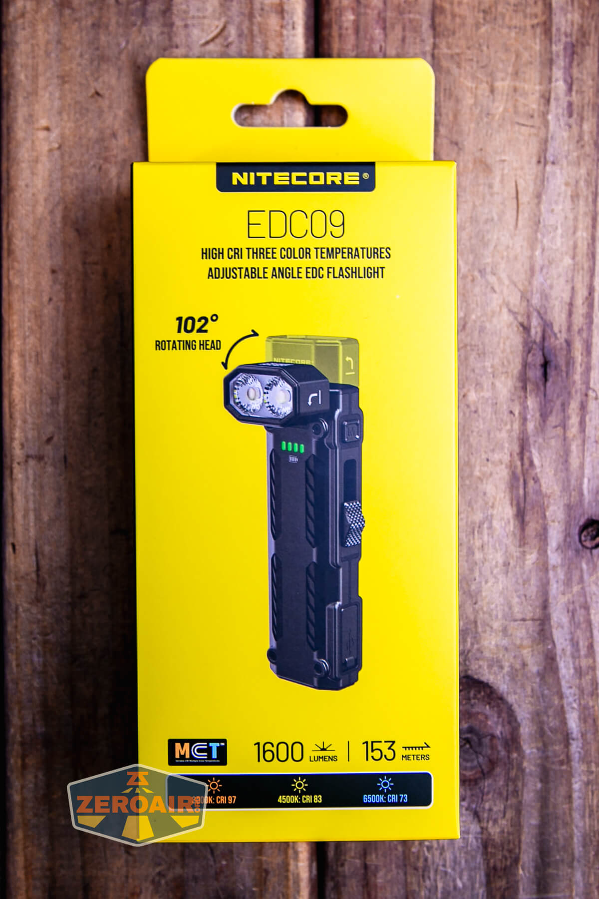 A yellow box for the Nitecore EDC09 flashlight is shown on a wooden surface. The box highlights features such as 102° rotating head, 1600 lumens, 153 meters range, and three color temperatures.