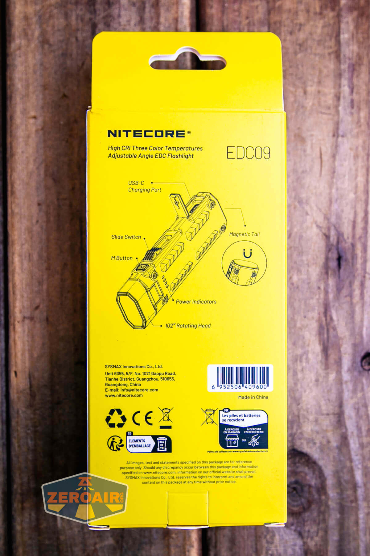 Back of a yellow Nitecore EDC09 flashlight box showing product features, labeled diagram of the flashlight, QR codes, barcodes, and company information, placed on a wooden surface.