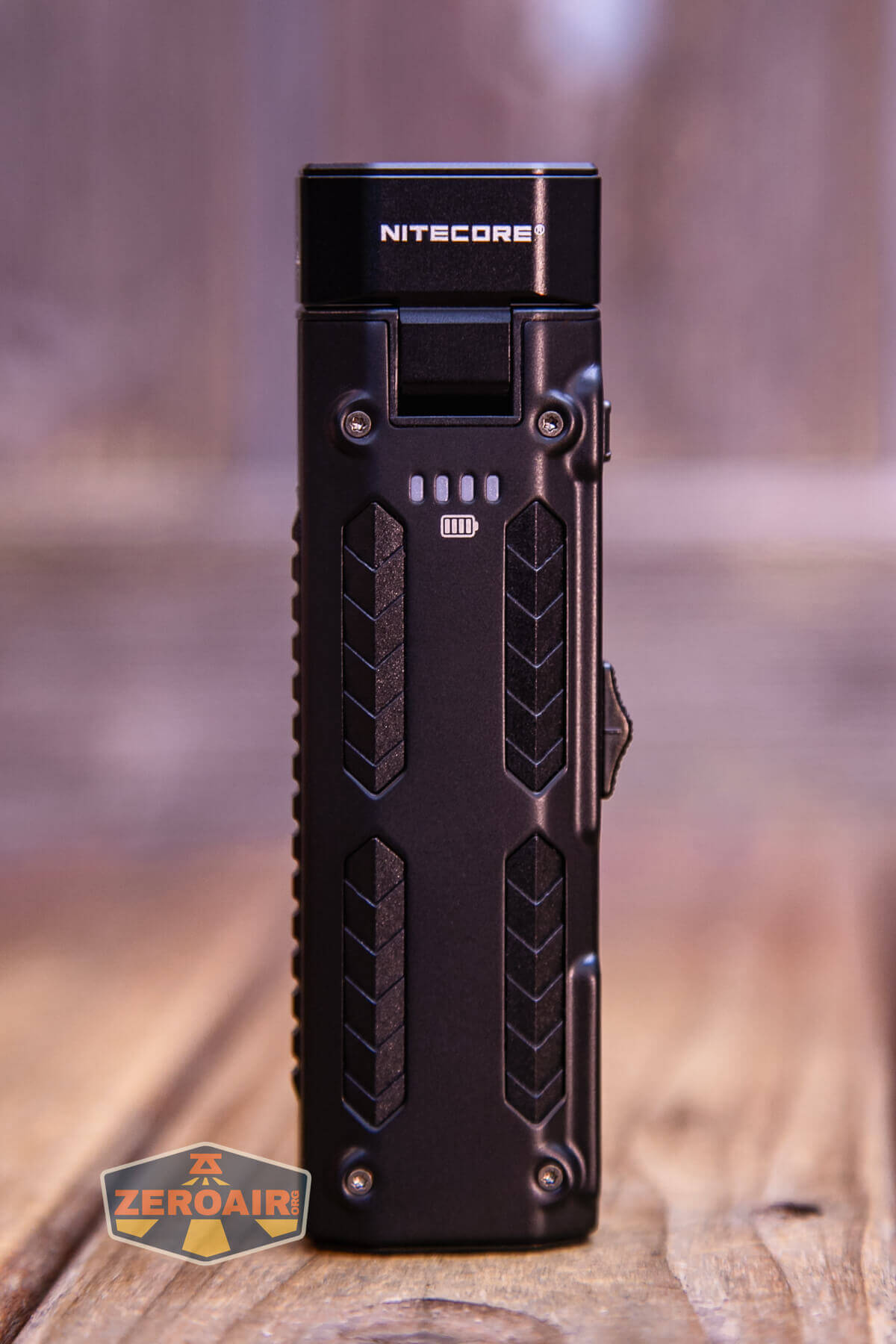 A black Nitecore device with a rectangular shape stands upright on a wooden surface. The front features a battery indicator and textured grip patterns. The background is softly blurred.
