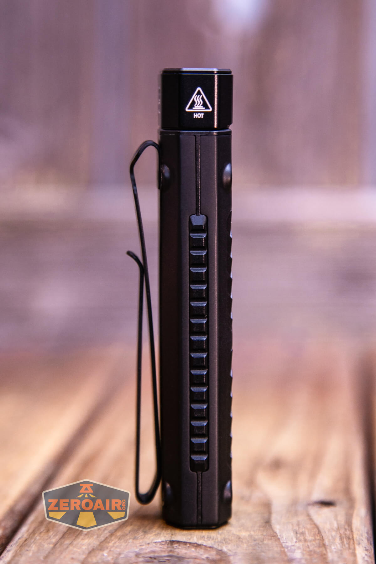 A slim, black flashlight stands vertically on a wooden surface with a clip attached to its side. The top is labeled HOT with a warning symbol. The background is softly blurred. A ZeroAir logo is in the bottom left corner.