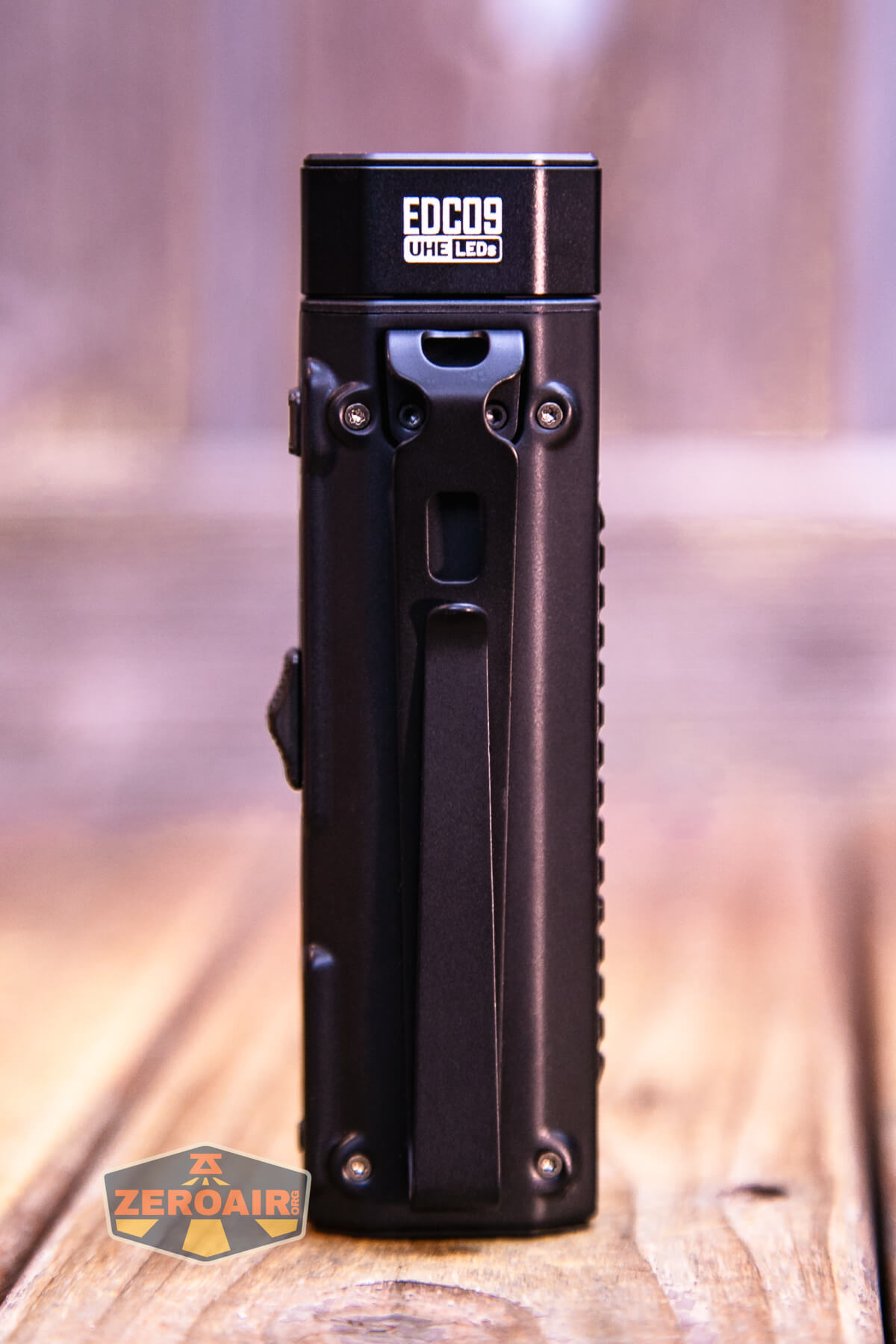 A black rectangular flashlight labeled EDC09 VHE LE Pro stands upright on a wooden surface, with a metal pocket clip and several screws visible on the body.
