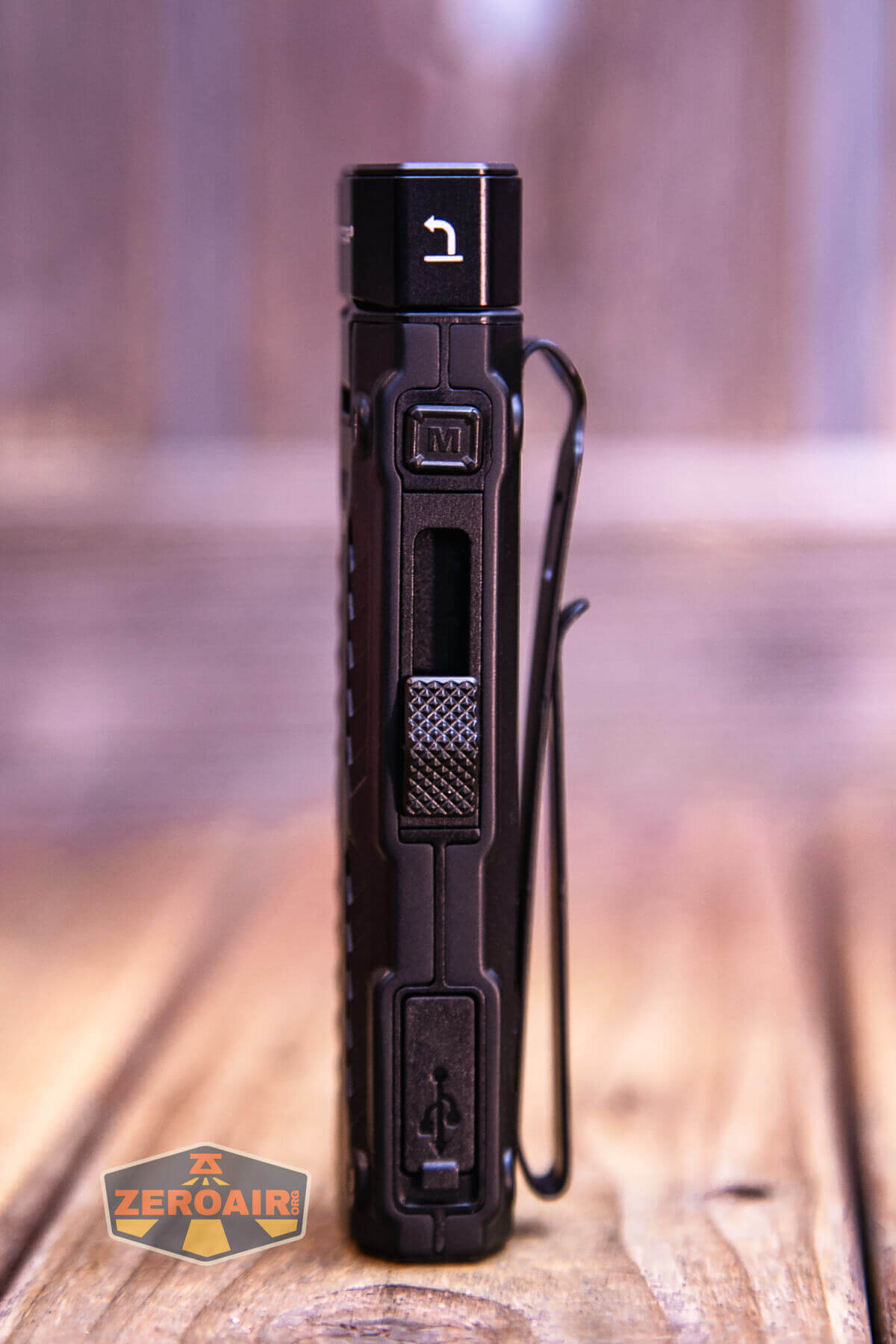 A black, rectangular flashlight with a metal clip is standing upright on a wooden surface. The flashlight has a button, a textured slider, and a visible charging port cover. A ZeroAir watermark is in the lower left corner.