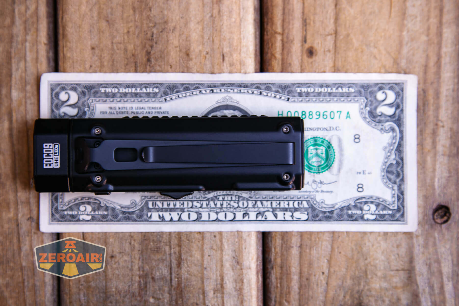 A black OTF knife with a pocket clip rests on top of a U.S. two-dollar bill placed on a wooden surface. An orange and gray ZeroAir logo is in the lower left corner of the image.