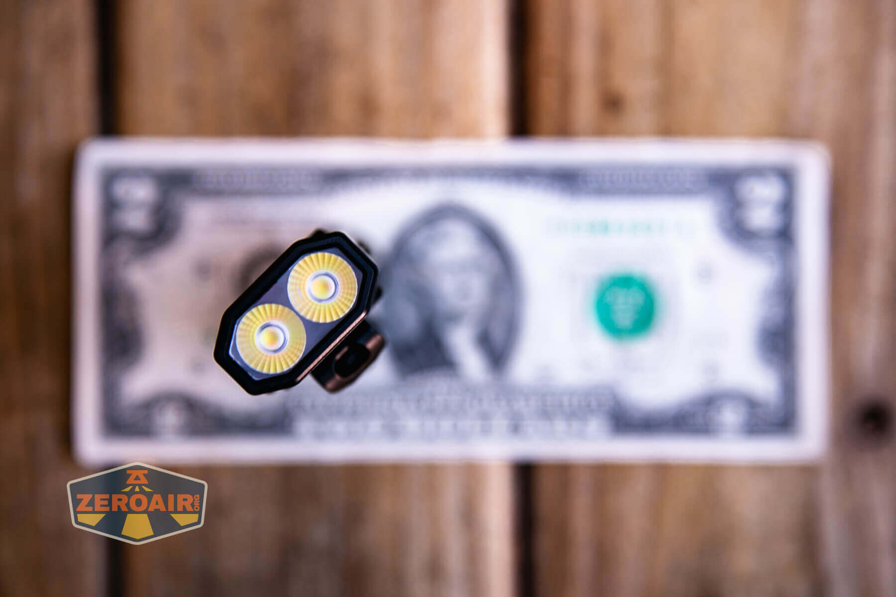 A close-up top view of a flashlight with two LED bulbs, positioned above a blurred U.S. two-dollar bill on a wooden surface. The ZeroAir logo is visible in the bottom left corner.