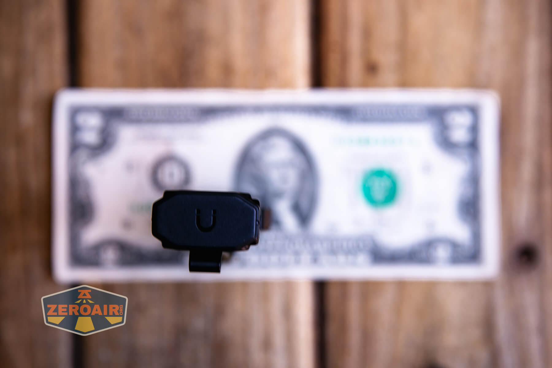 A handgun is placed upright on a wooden surface, aimed at the viewer, with a U-shaped rear sight in focus. A US one-dollar bill lies flat beneath the gun, and a ZeroAir logo is visible in the lower left corner.