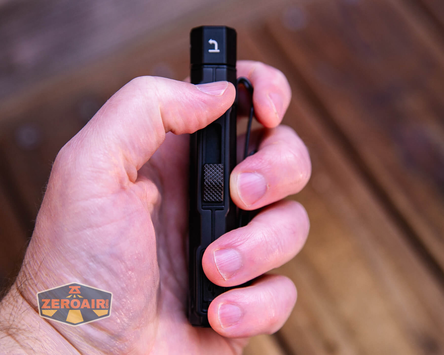 A hand holding a small black flashlight with a clip, positioned vertically. The thumb rests on a textured button. A wooden surface and a ZeroAir logo are visible in the background.