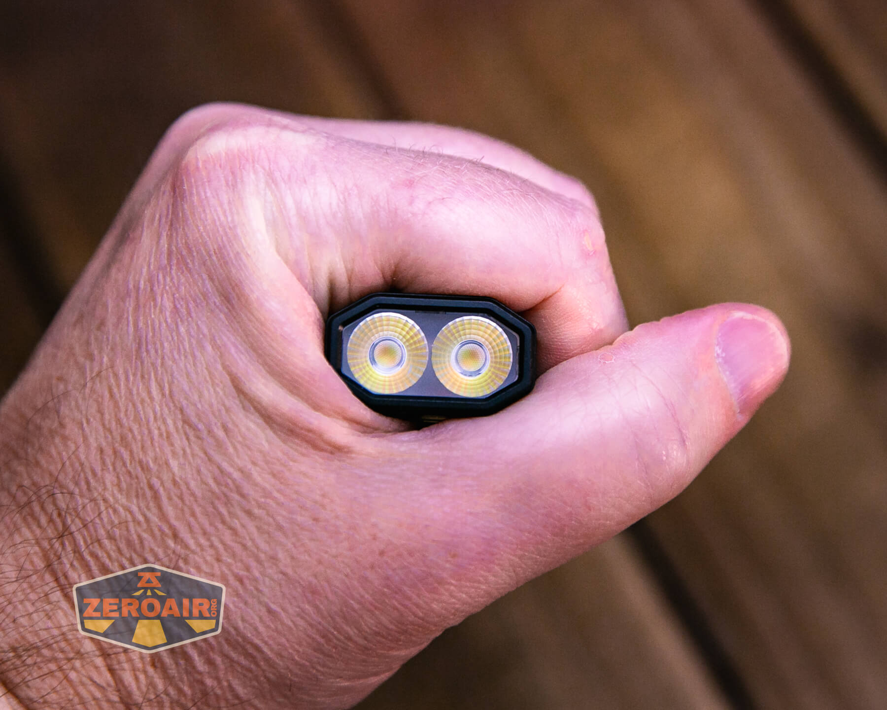A hand holding a rectangular flashlight with two circular LED lights visible at the end. The hand grips the flashlight tightly, and a ZeroAir logo is seen in the bottom left corner.