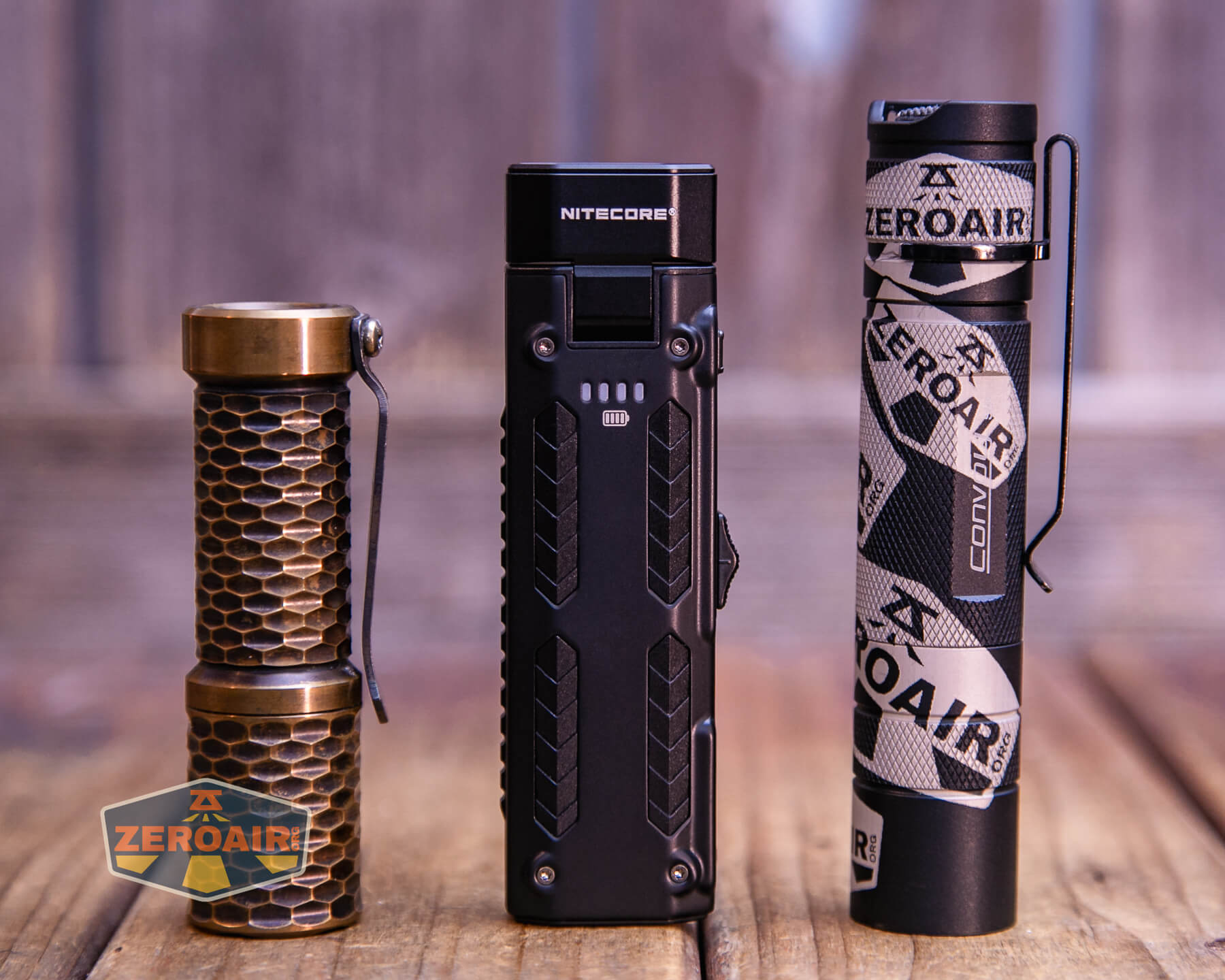 Three flashlights are standing upright on a wooden surface. The left flashlight has a bronze honeycomb design, the middle one is black and rectangular labeled NITECORE, and the right one is black with white branding and a clip.