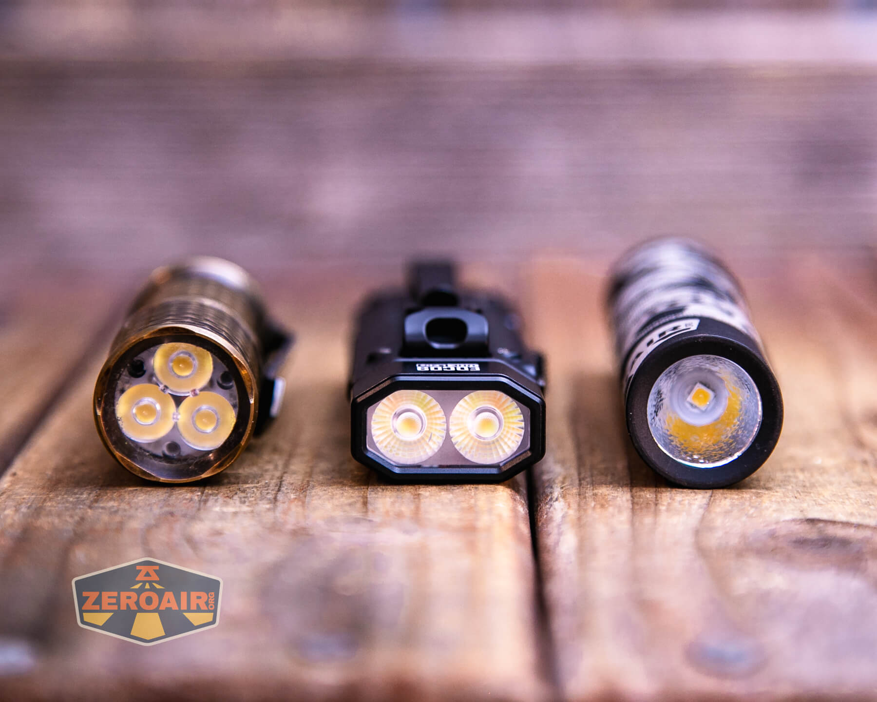 Three flashlights are placed upright on a wooden surface, showing their LED arrays. The flashlight in the center has rectangular dual LEDs, while the ones on the left and right have circular LED configurations.