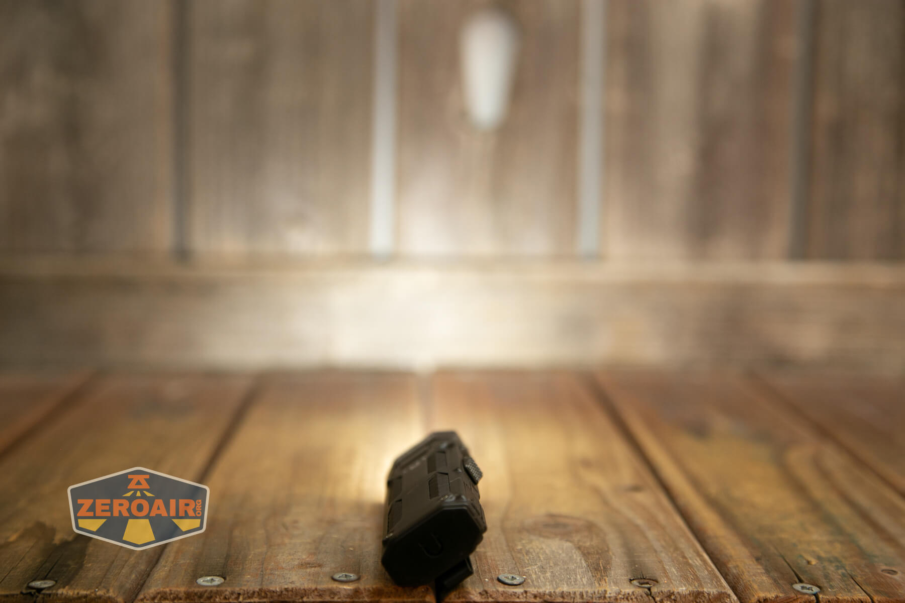 A black flashlight lies on a wooden surface with a blurred wooden wall in the background. The lower left corner features a ZeroAir! logo.