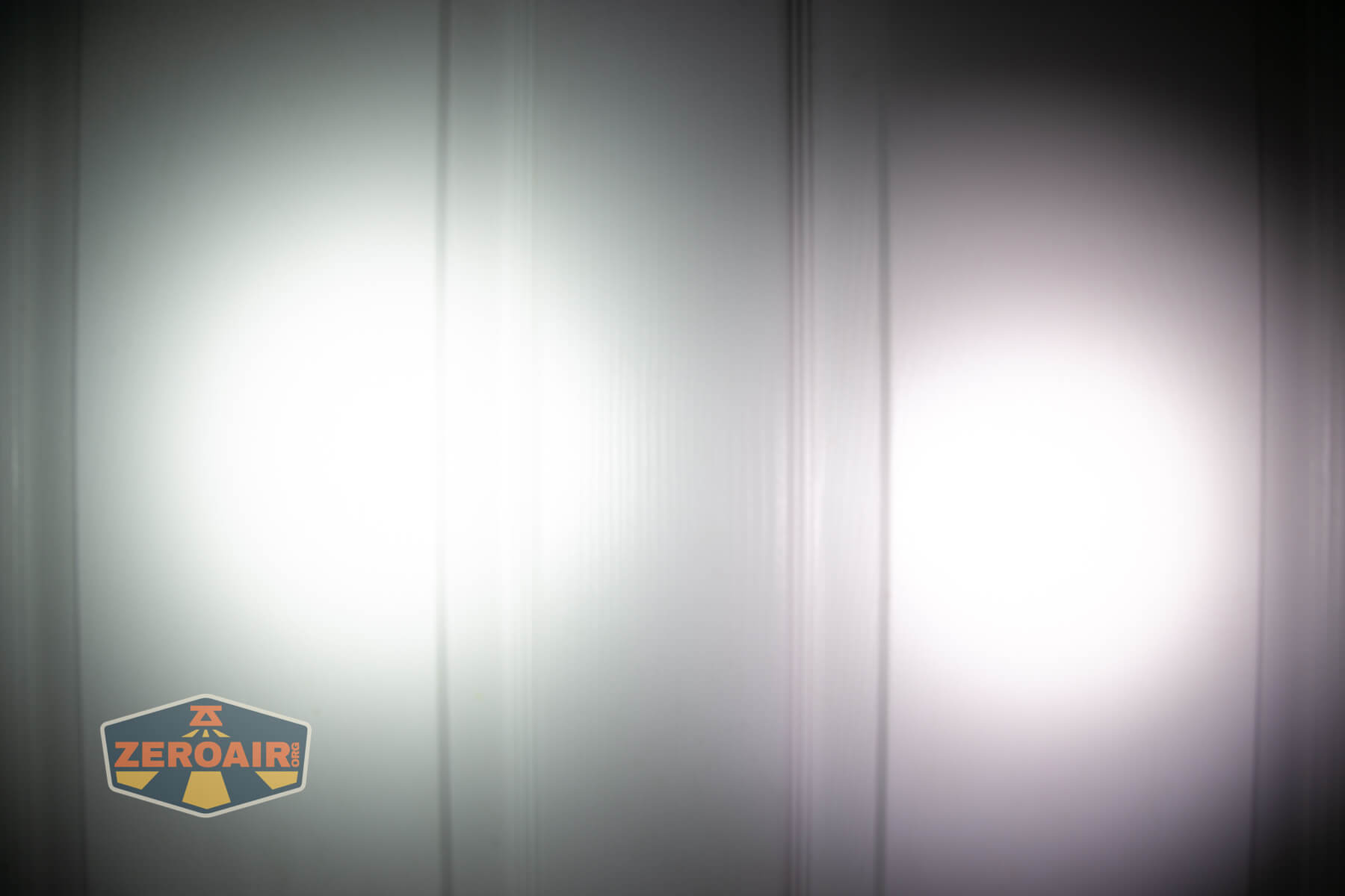 Two bright flashlight beams shine on a white paneled surface, creating circular patterns of light. A logo in the bottom left corner reads ZeroAir.