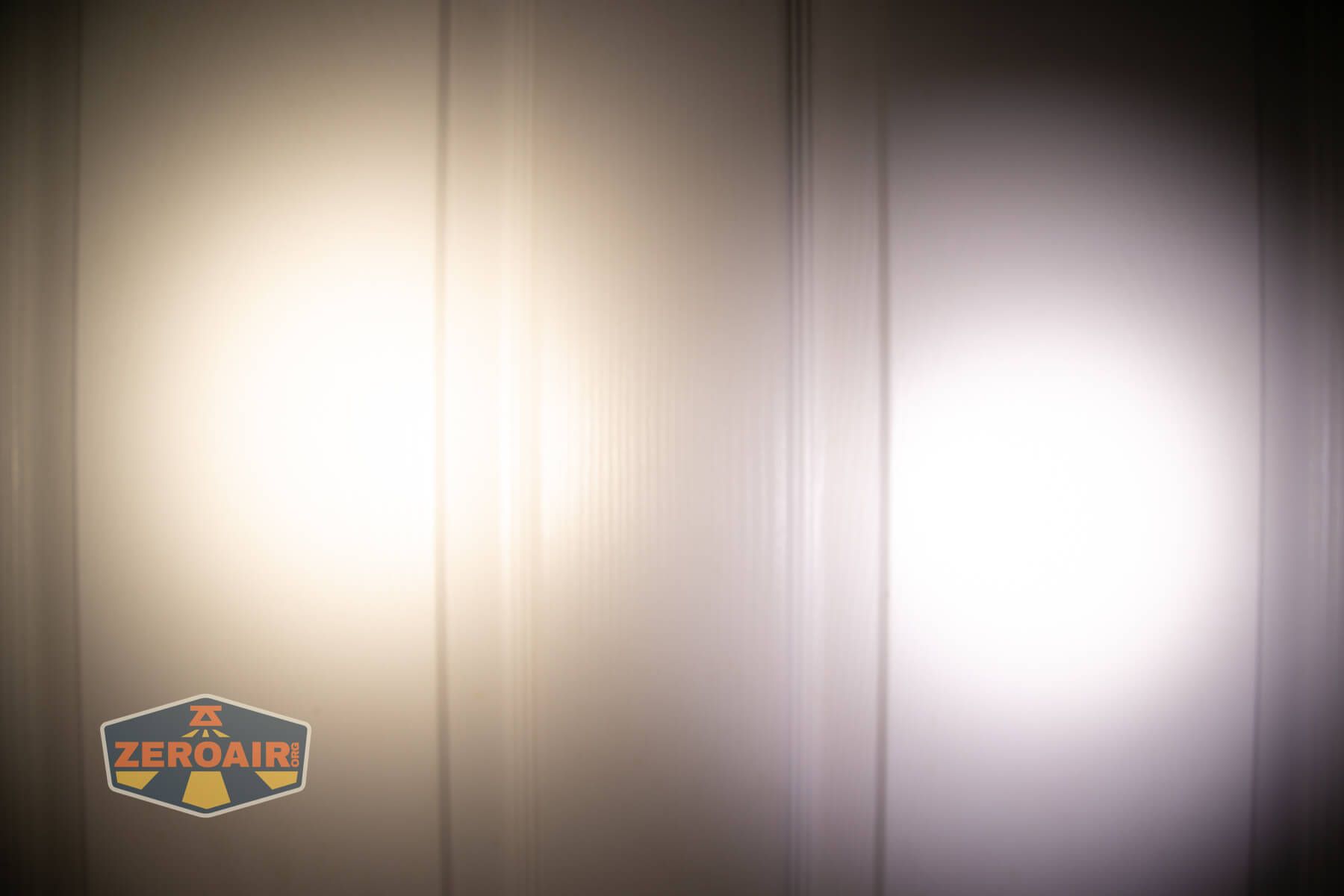 Two circular flashlight beams, one warm white (left) and one cool white (right), shine on a vertically paneled surface. Both beams have defined centers with soft edges. A ZeroAir logo is seen in the lower left corner.