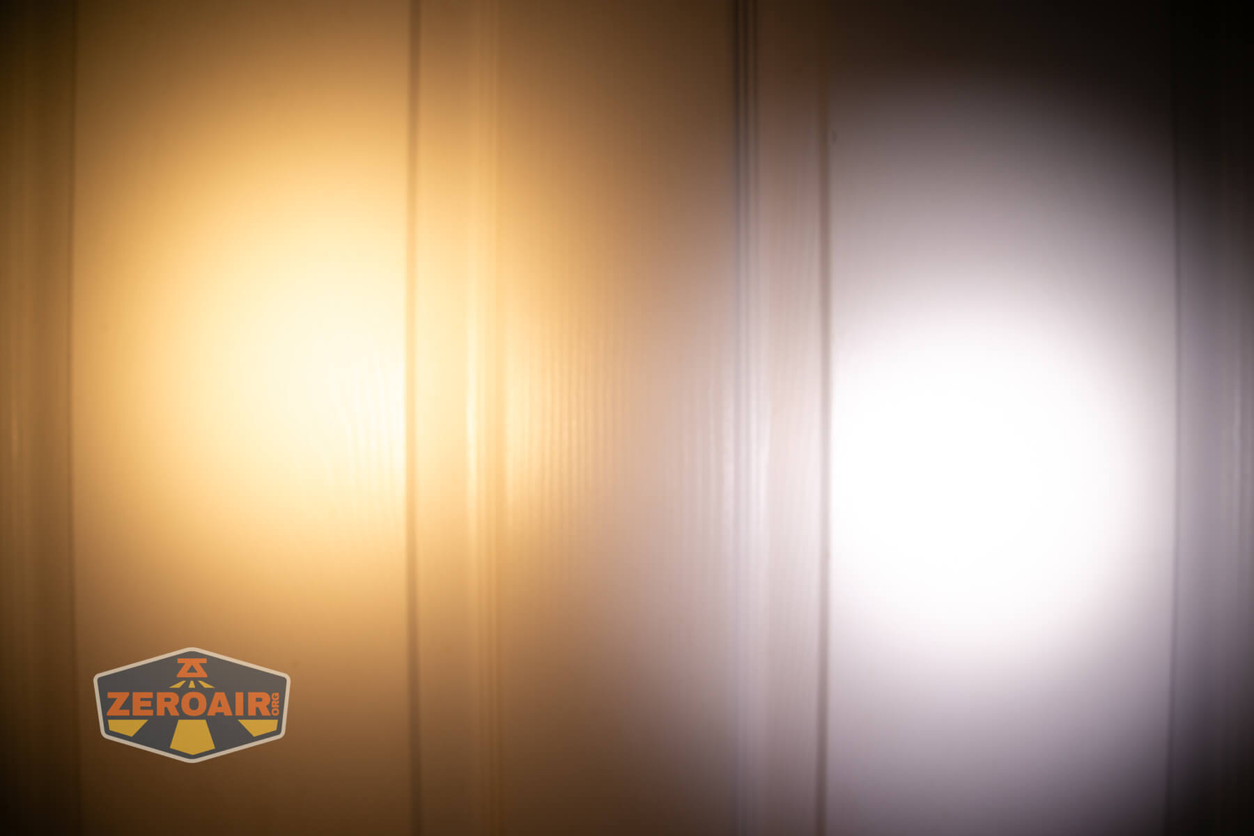 Two beams of light, one warm yellow and one cool white, are projected onto a vertical paneled surface. A Zeroair! logo is visible in the lower left corner of the image.