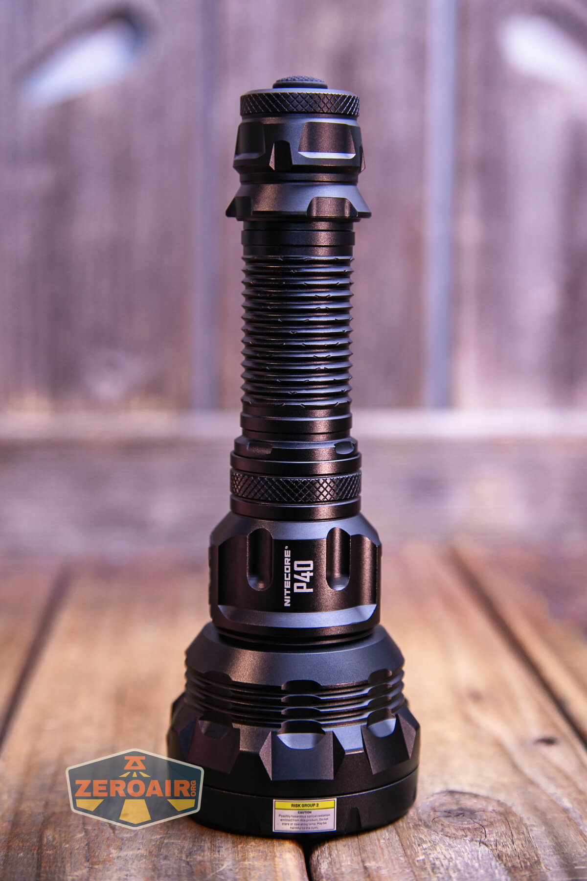 A black, heavy-duty flashlight stands upright on a wooden surface with a blurred wood background. The flashlight has a textured handle and a wide head. A “ZeroAir” logo is visible in the lower-left corner.