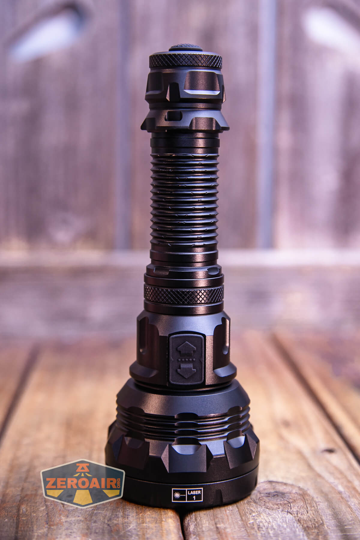A black tactical flashlight with a textured grip and a wide base stands upright on a wooden surface. There is a ZeroAir logo in the bottom left corner. The background is blurred wood planks.