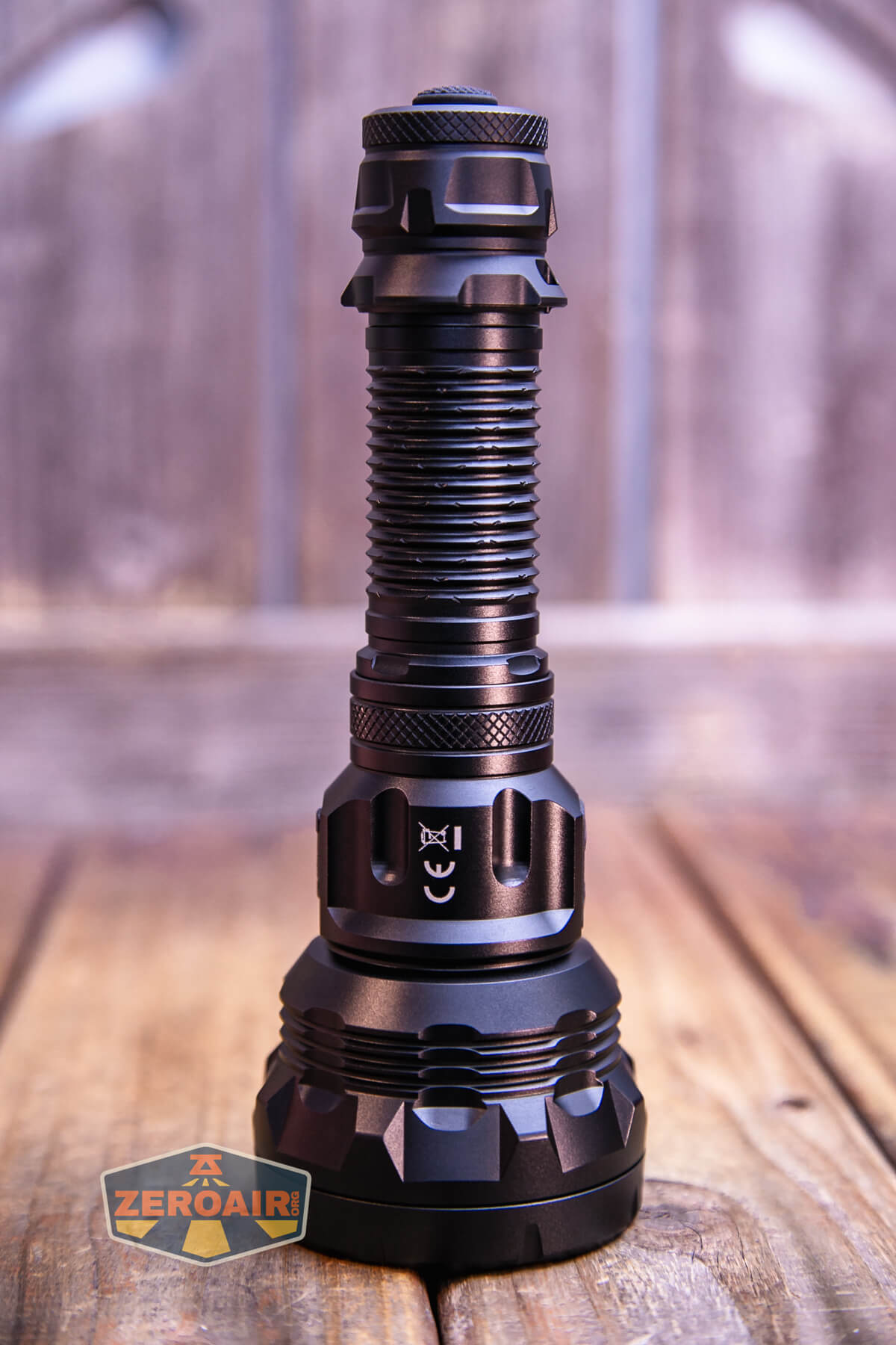 A black, heavy-duty flashlight stands upright on a wooden surface with a blurred wood panel background. The ZeroAir logo is visible in the lower left corner.