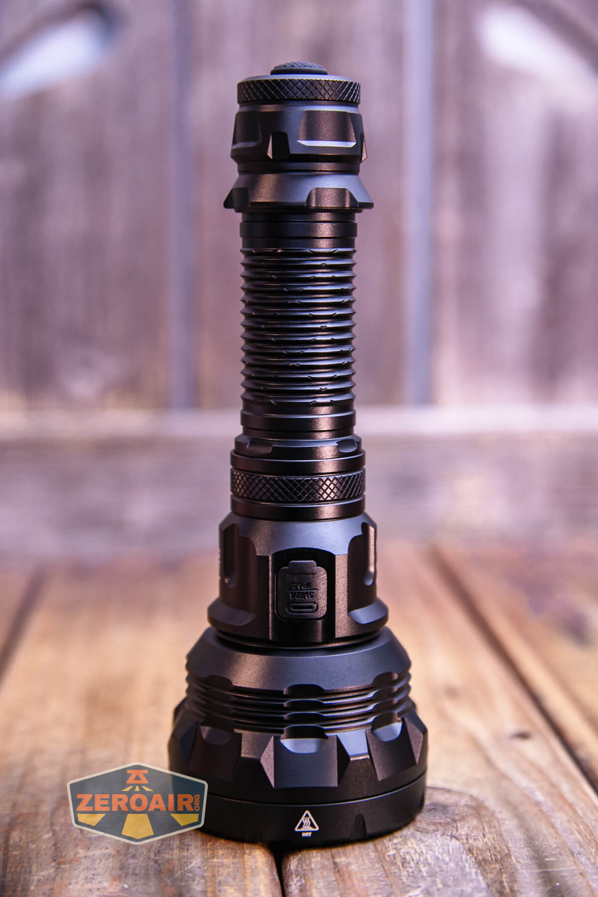 A black, heavy-duty flashlight stands upright on a wooden surface. The body has ridged and textured details, and a ZeroAir logo appears in the lower left corner. The background is blurred wood.
