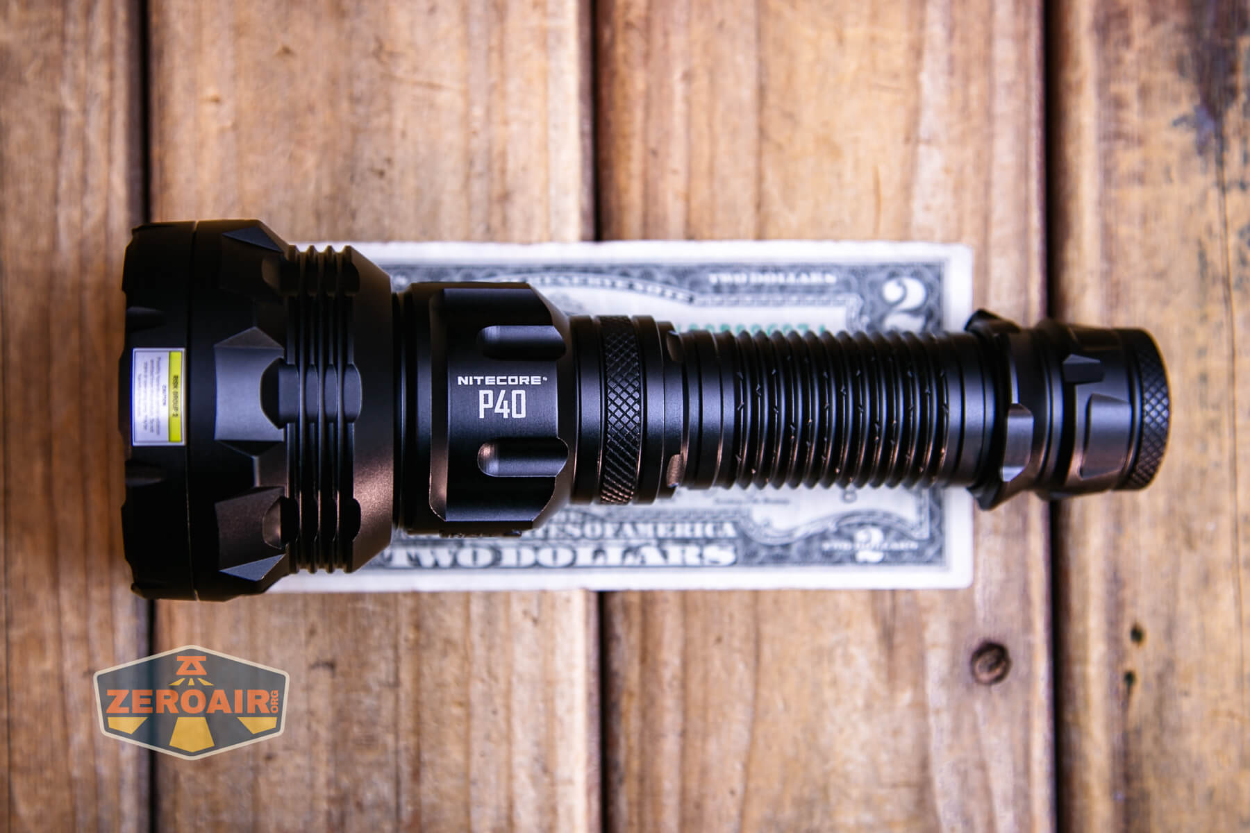 A Nitecore P40 flashlight is placed horizontally on top of a two-dollar bill on a wooden surface. A ZeroAir logo appears in the bottom left corner of the image.