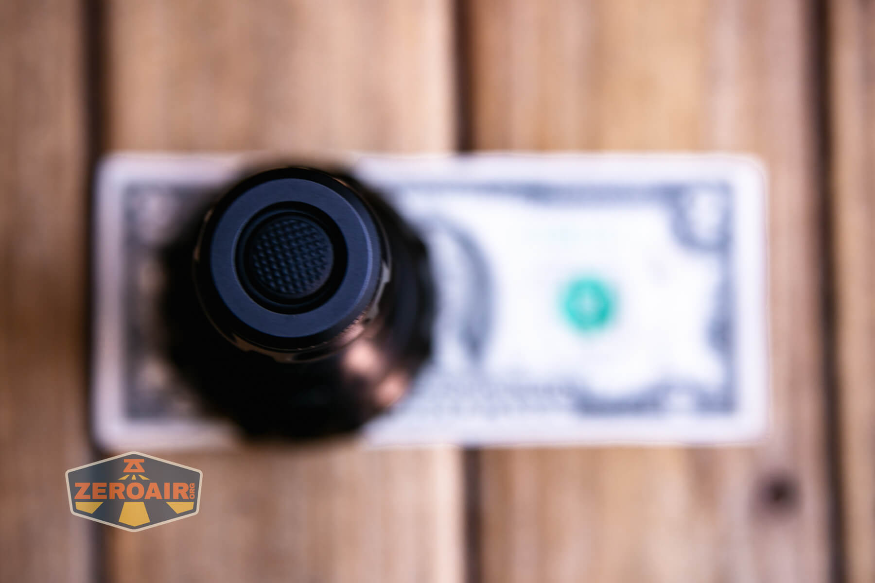 A close-up, top-down view of a black flashlight standing upright on a blurred U.S. one-dollar bill, placed on a wooden surface. A ZeroAir logo is visible in the bottom left corner.