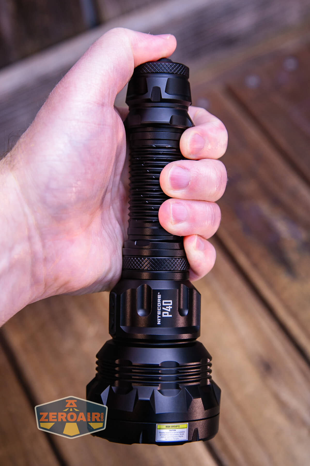 A hand holding a large black flashlight vertically over a wooden surface; the flashlight has textured grip sections and visible ridges, with a ZeroAir logo in the lower left corner.