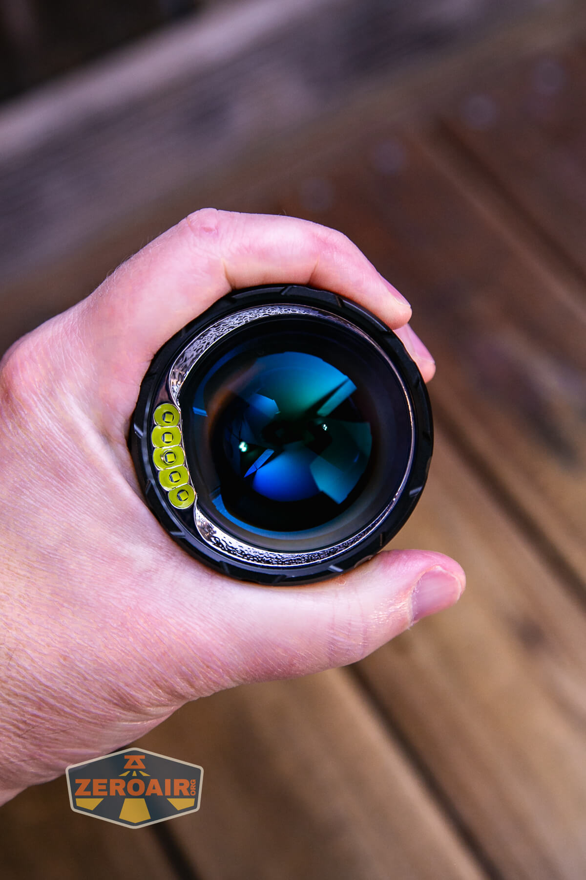A hand holding a flashlight, viewed from the lens end. The flashlight reflects blue and green hues, and the logo ZEROAIR appears on the bottom left. The background is wooden.