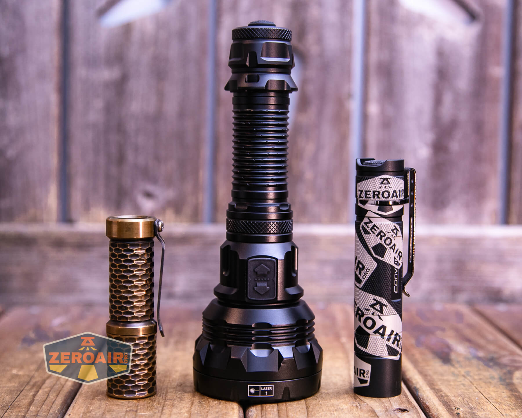 Three flashlights of varying sizes and designs are standing upright on a wooden surface in front of a wooden background. The flashlight on the left has a bronze honeycomb pattern, the middle one is large and black, and the right one is black and white with a pattern.
