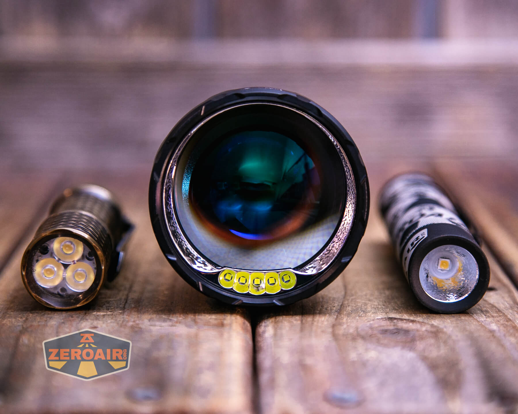 A close-up of three flashlight components on a wooden surface: a flashlight head in the center, a battery on the right, and an LED module on the left. The ZeroAir logo is visible in the bottom left corner.