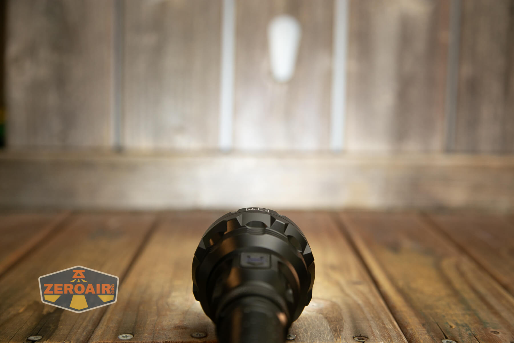 A flashlight is turned on and placed on a wooden surface, shining a beam of light onto a wooden wall. The ZeroAir logo is in the lower left corner of the image.