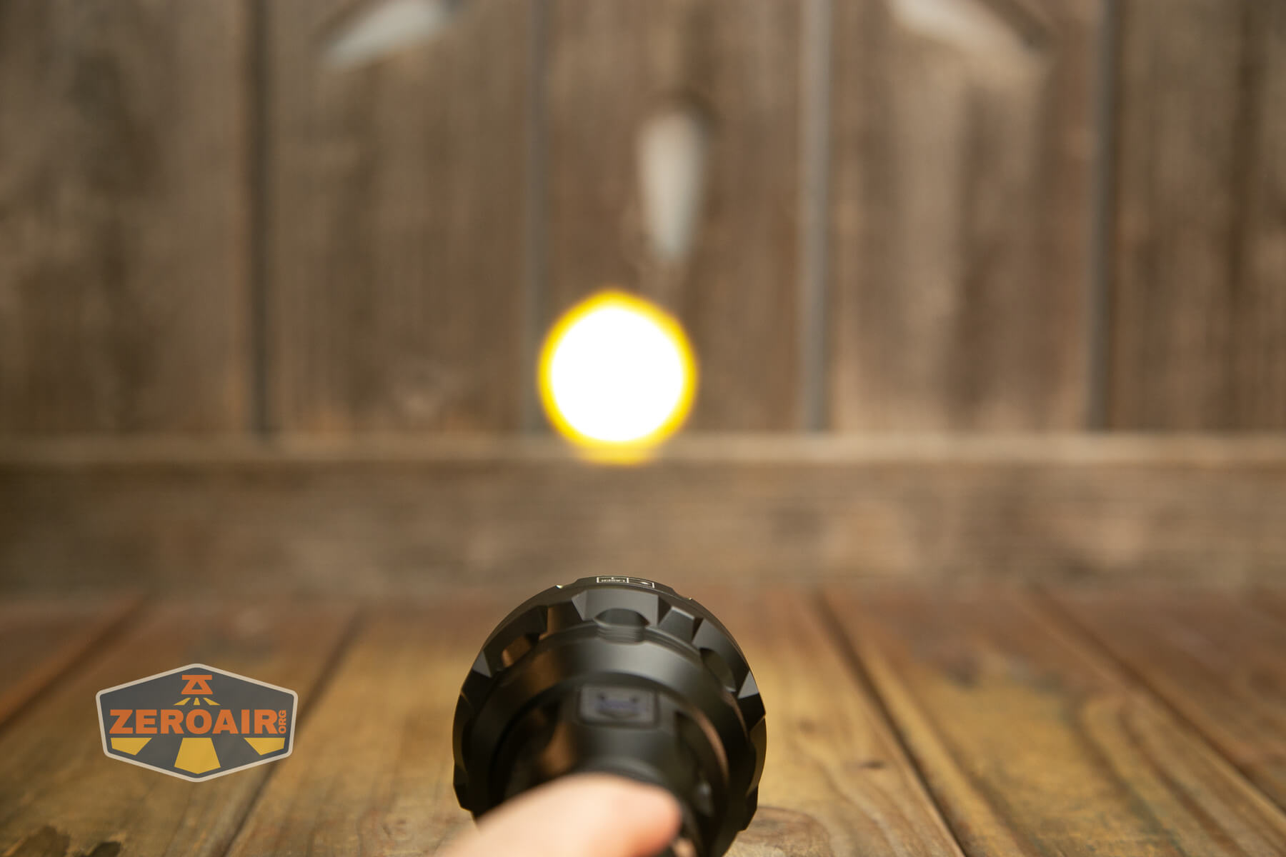 A hand holds a flashlight pointed at a wooden fence, illuminating a bright circular spot on the fence. Wooden floorboards are visible, and a logo reading ZEROAIR is in the lower left corner.