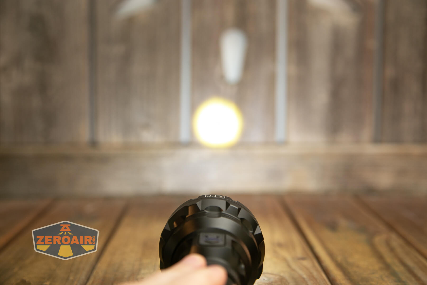 A hand holds a flashlight, shining a bright circular beam onto a wooden wall with vertical panels. The ZeroAir logo is visible in the lower left corner of the image.