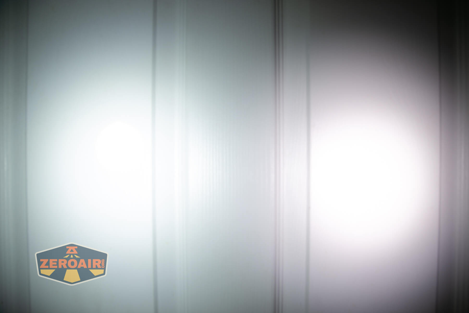 Two white flashlight beams shine on a white paneled wall, creating overlapping bright circular spots. A logo reading ZeroAir appears in the lower left corner of the image.