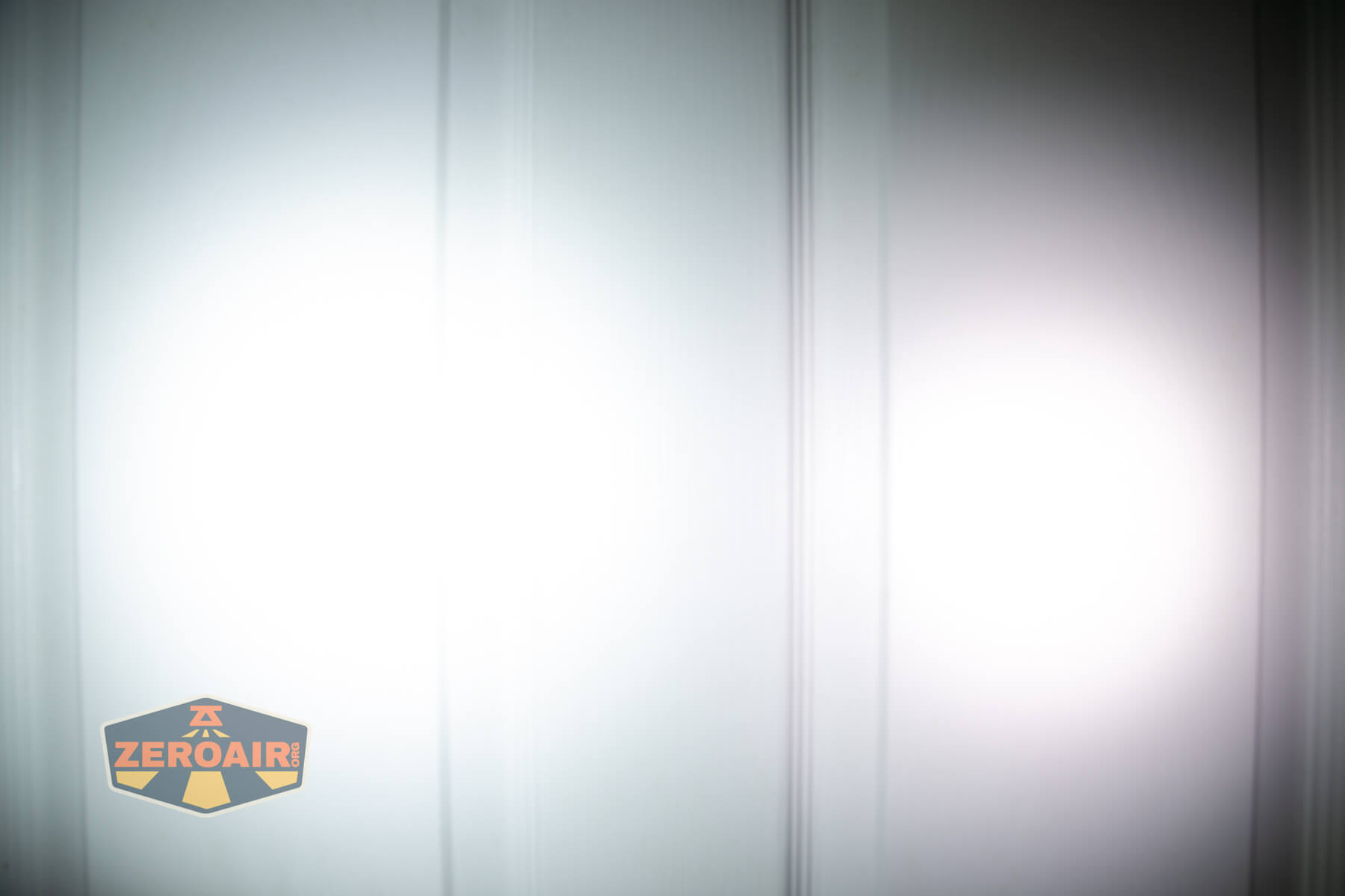 Two overlapping bright white spots of light are projected onto a smooth white surface, possibly a door. The image includes a ZeroAir logo in the lower left corner.