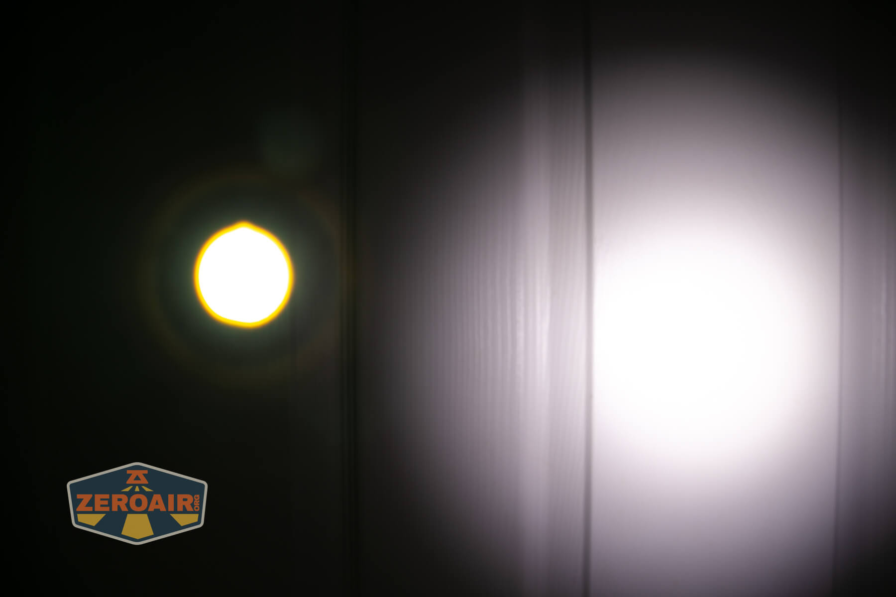 A bright yellowish circular light and a larger white spotlight are projected onto a dark, vertical-paneled surface. The ZeroAir logo is visible in the lower left corner of the image.