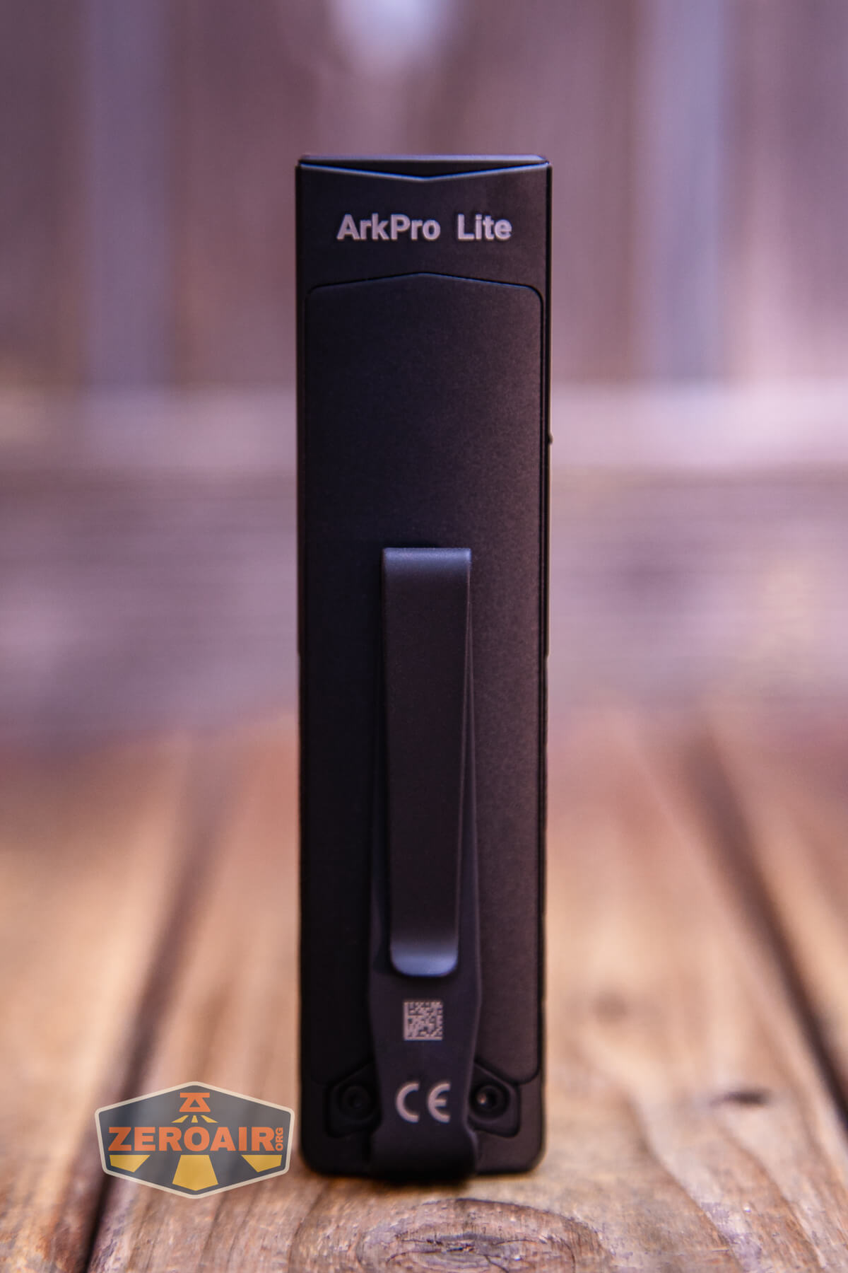 A black rectangular device labeled ArkPro Lite is standing upright on a wooden surface. The device features a clip and certification markings on its front. The background is blurred.