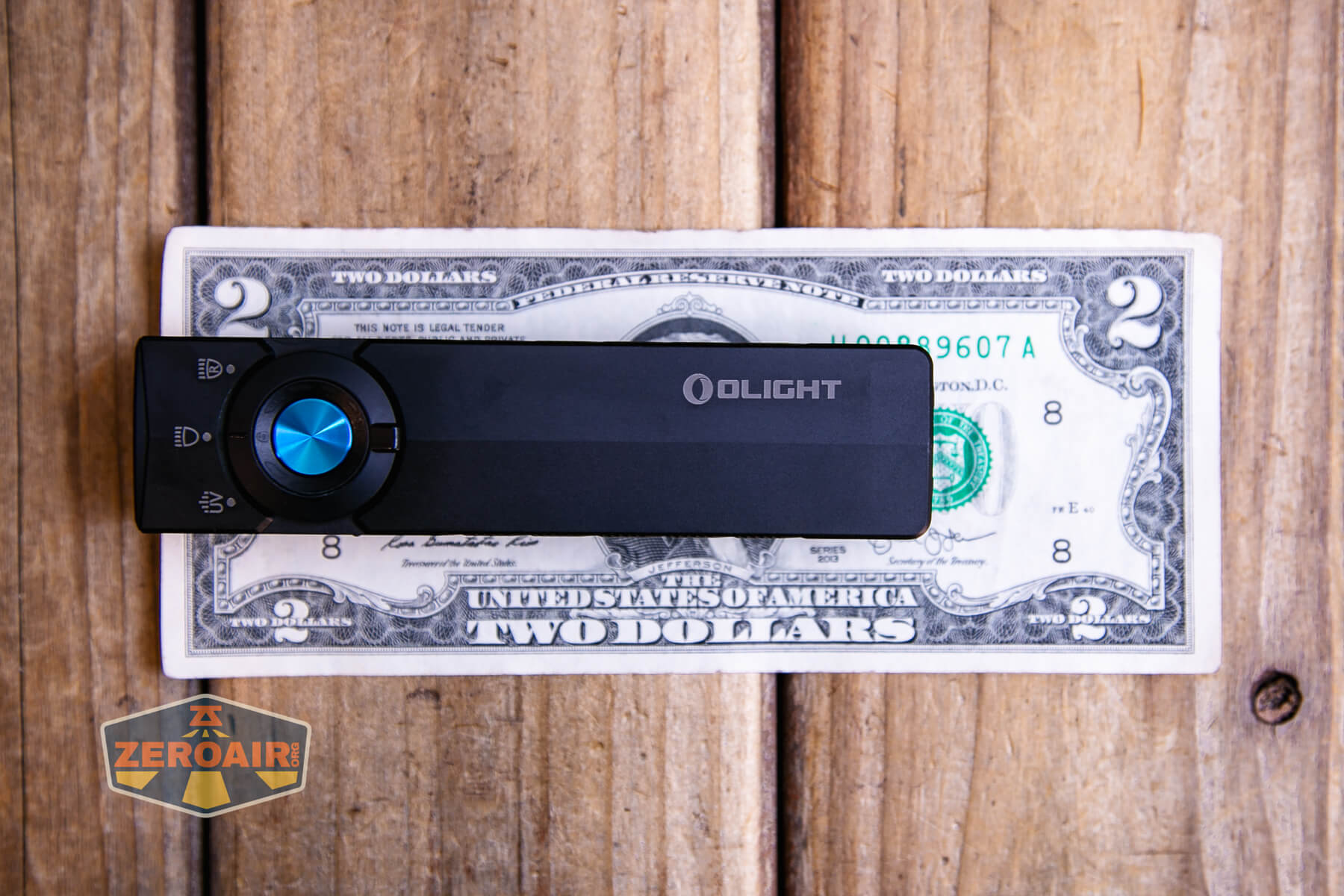 A rectangular black Olight device with a blue circular dial is placed on top of a two-dollar bill, all set on a wooden surface. A sticker showing the ZeroAir logo is in the bottom left corner.