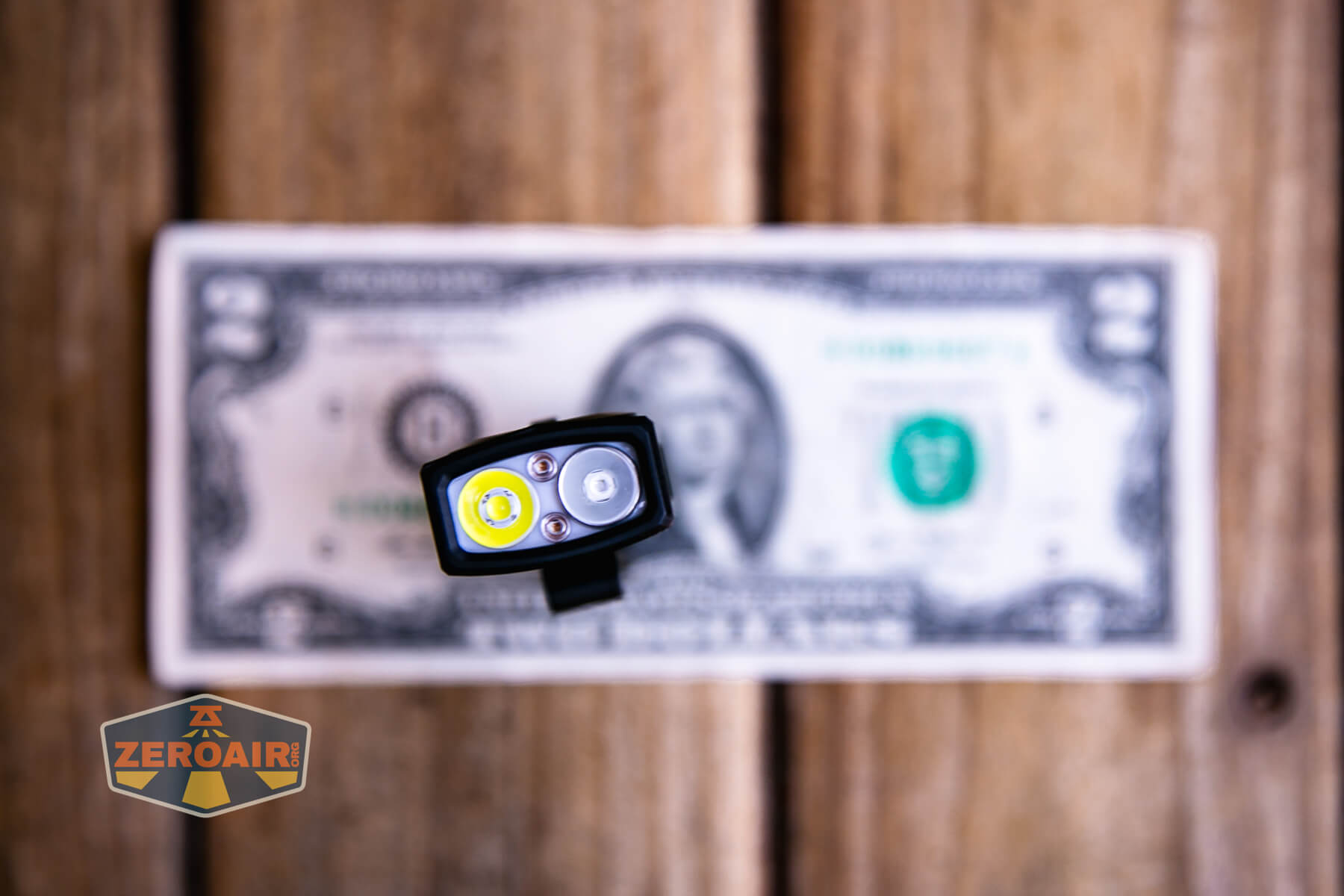 A small flashlight shown from above, displaying its two LED bulbs, is positioned in front of a two-dollar bill on a wooden surface. A ZeroAir logo appears in the bottom left corner.