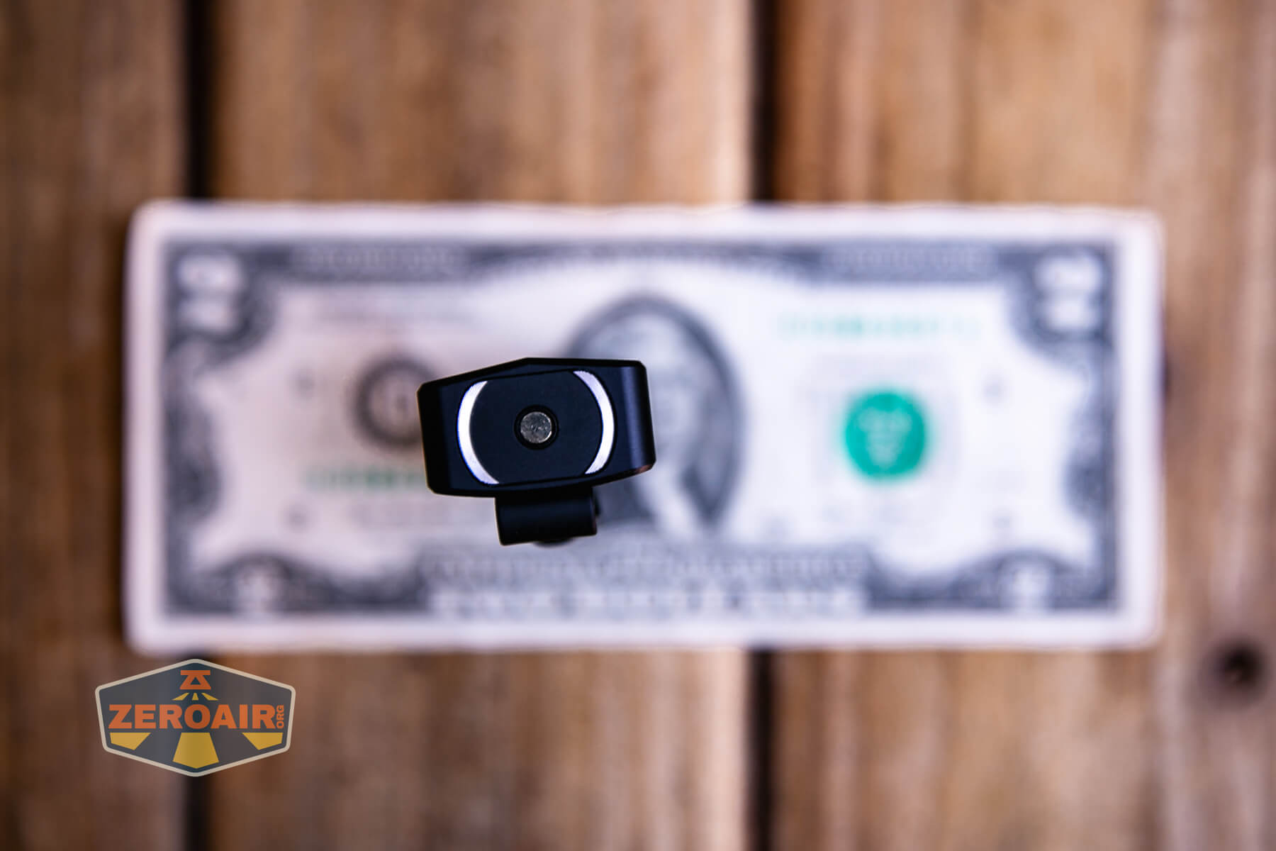 A black flashlight standing vertically on top of a U.S. one-dollar bill, placed on a wooden surface. The flashlights lens faces the camera. A logo with the word ZEROAIR! is in the bottom left corner.