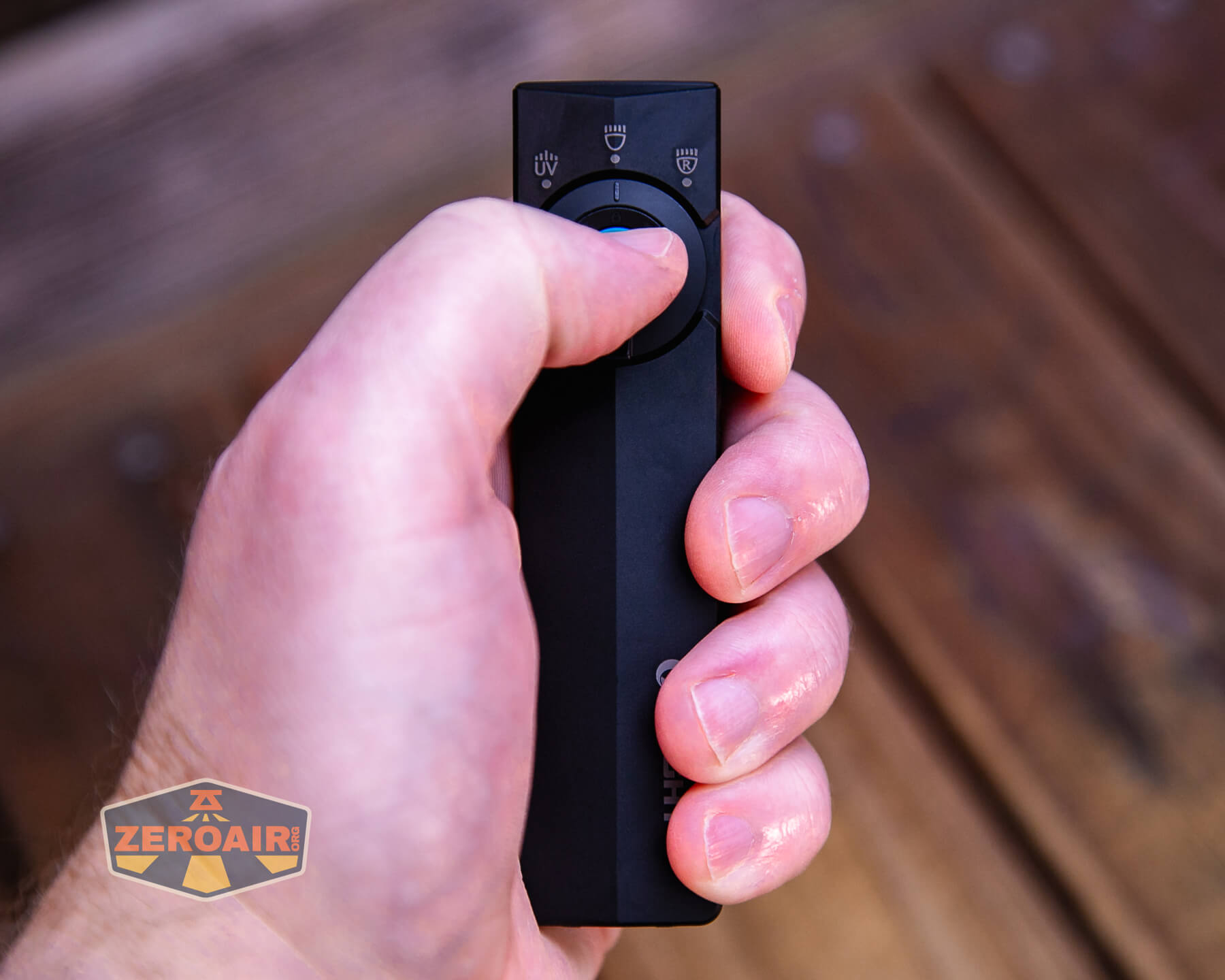 A hand holding a black remote control, pressing a central button. The background shows a wooden surface, and a “Zeroair” logo is visible in the bottom left corner.